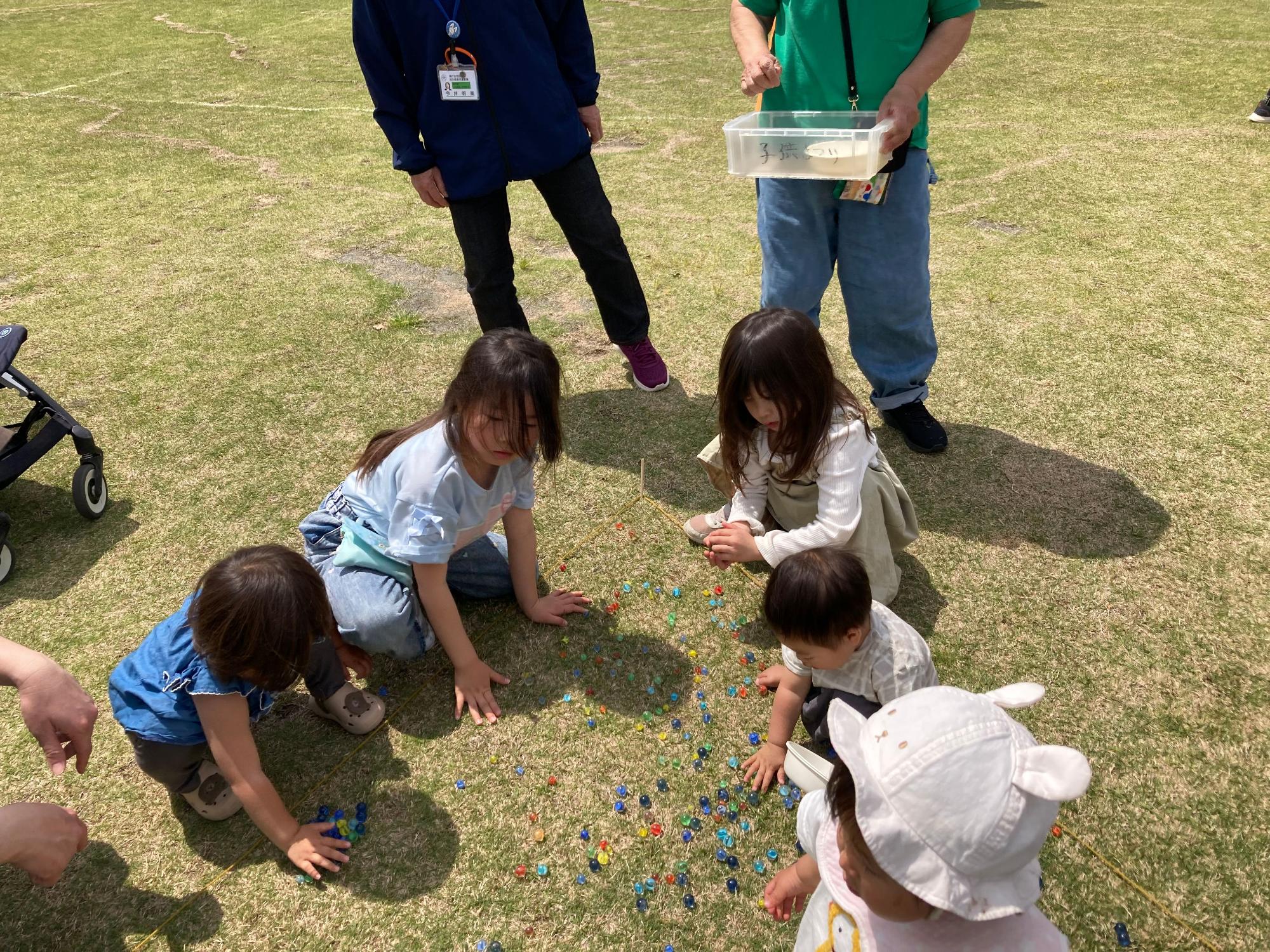 緑の芝生の上にたくさんのビー玉が散らばっており、それを囲んで子供たちが、大人に見守られながら夢中になって遊んでいる写真