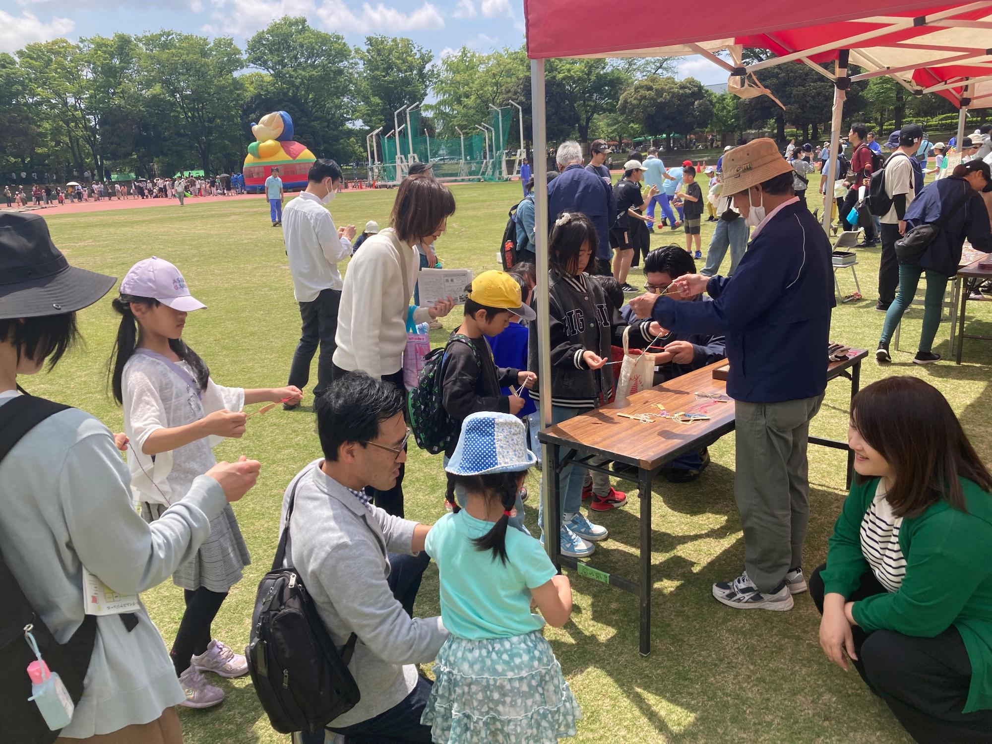 広場の芝生の上で、大人たちが子どもたちに昔の遊びを教えるイベントの様子を撮影した写真