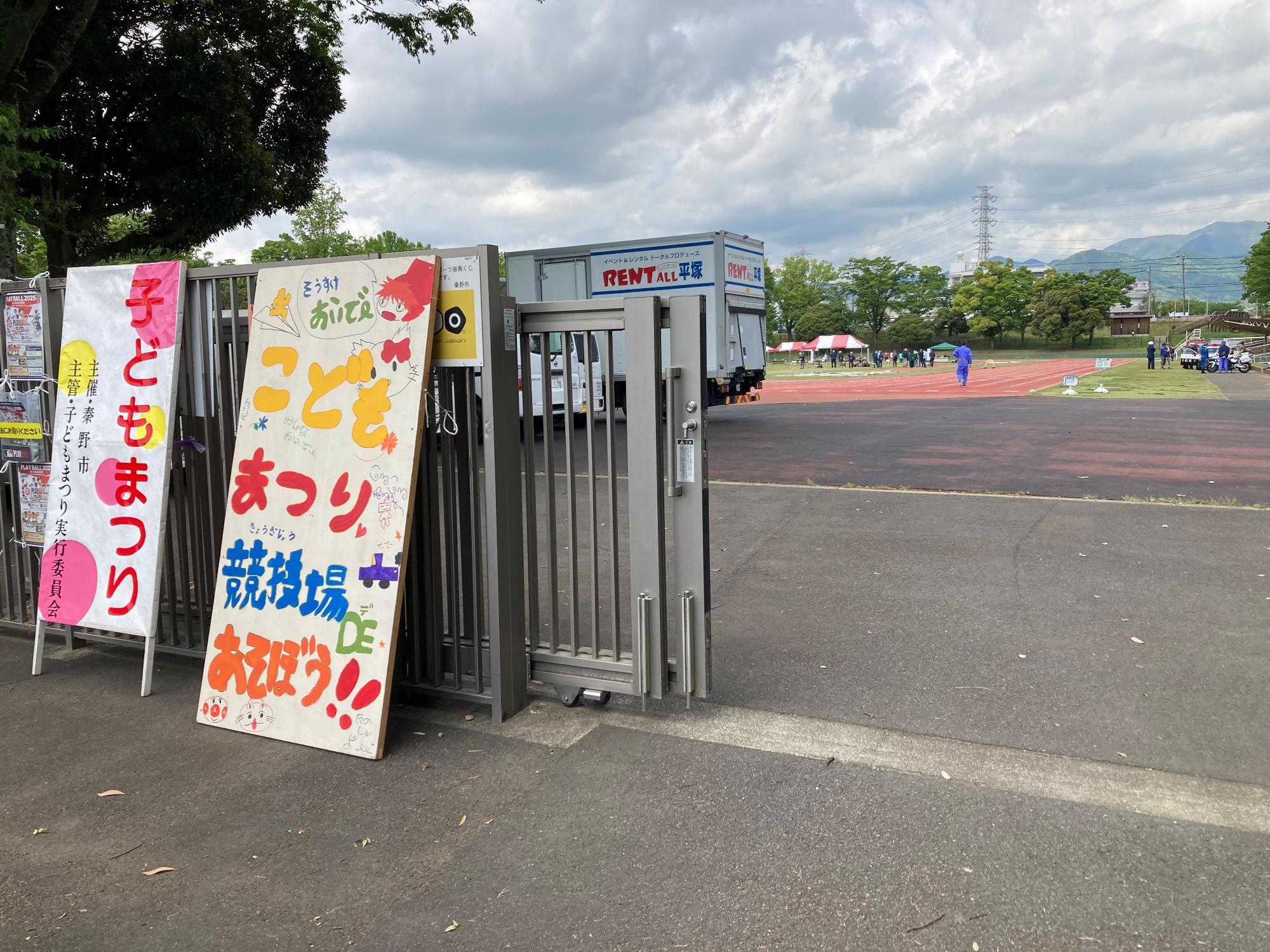 運動場の入り口に「子どもまつり」と書かれた看板が置かれ、トラックの向こうでは複数の人がイベントの準備をしている写真