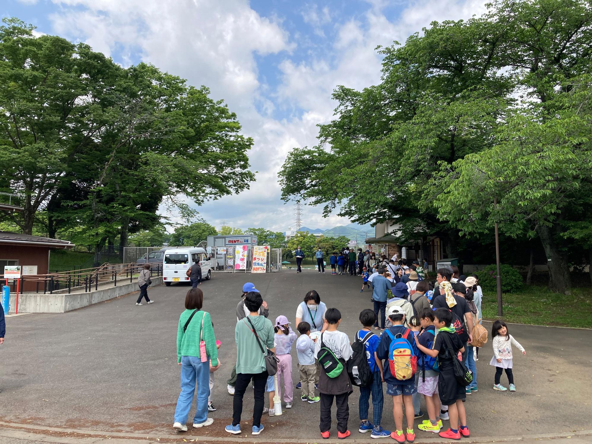 快晴の空のもと、公園の入り口に大勢の子供たちが集まっており、遠くにはイベントスタッフが準備をしている屋外写真