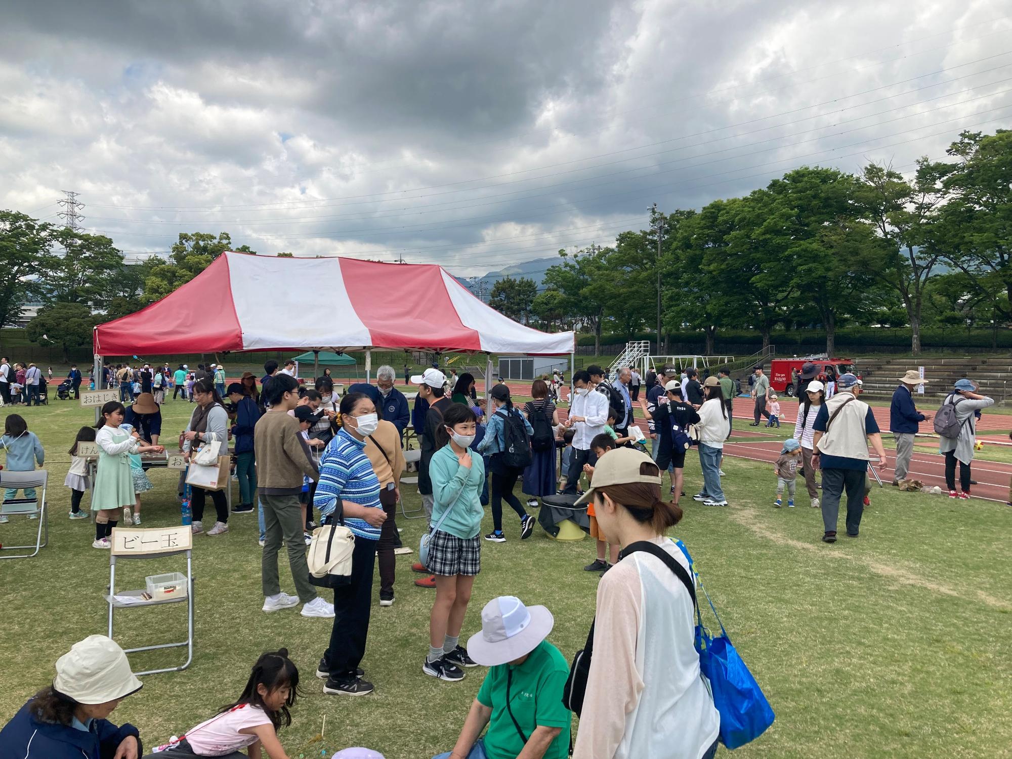 運動場の広場に設置された大きな紅白のテントの周りで、大勢の家族連れや子どもたちが集まり、様々なイベントを楽しんでいる賑やかな屋外写真
