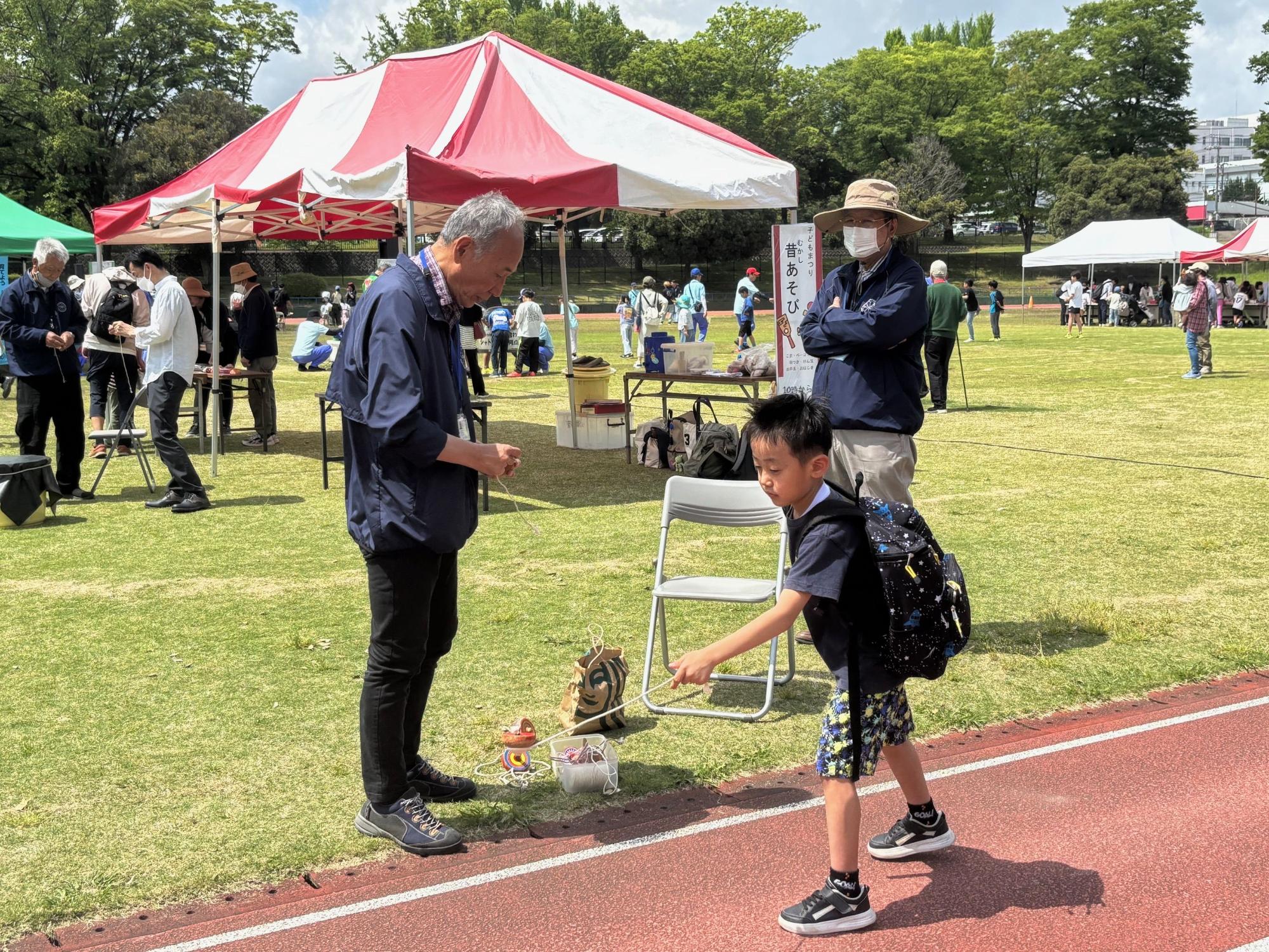 広い屋外広場のトラックと芝生の上で、大人と子供たちが、コマ回しのような昔の遊びを楽しんでおり、後ろにはイベント用のテントが複数設営されている写真