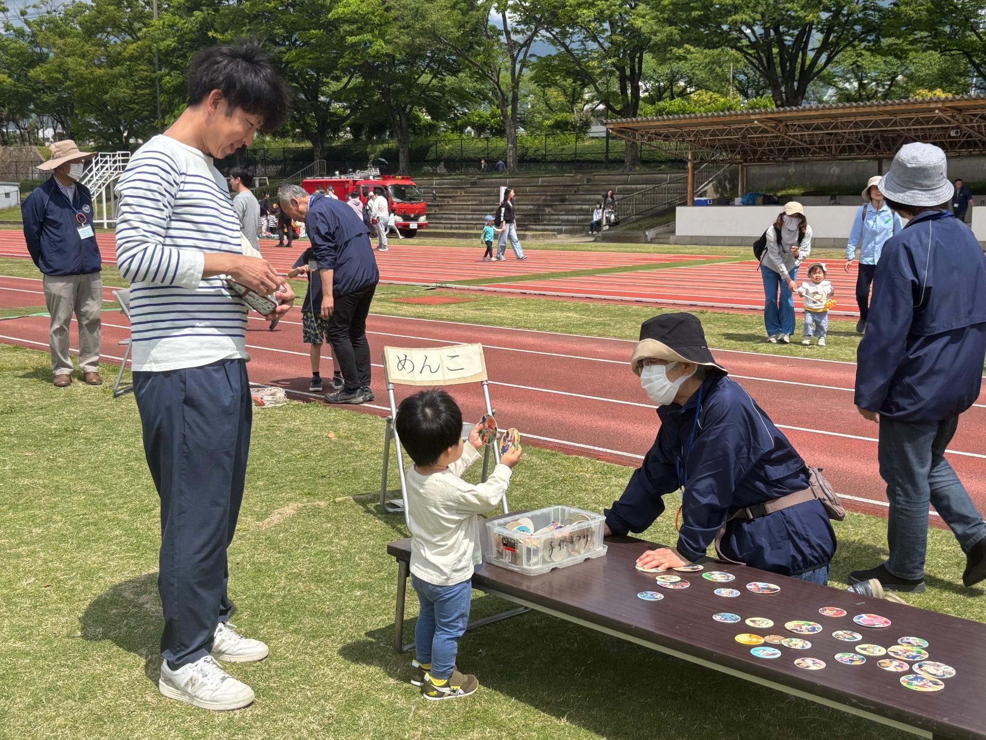 芝生と陸上競技用トラックのある広場で、親子がテーブルを囲んで「めんこ」遊びを楽しんでおり、周りには他の参加者もいる屋外イベントの様子の写真