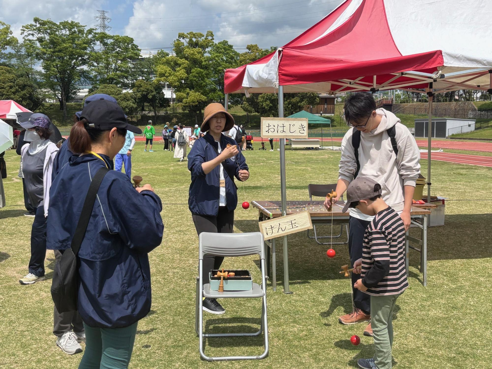 陸上競技場の芝生エリアで、赤いテントと「おはじき」や「けん玉」と書かれた看板が置かれたブースにて、大人が子どもたちに昔ながらの遊びを教えている屋外イベントの写真