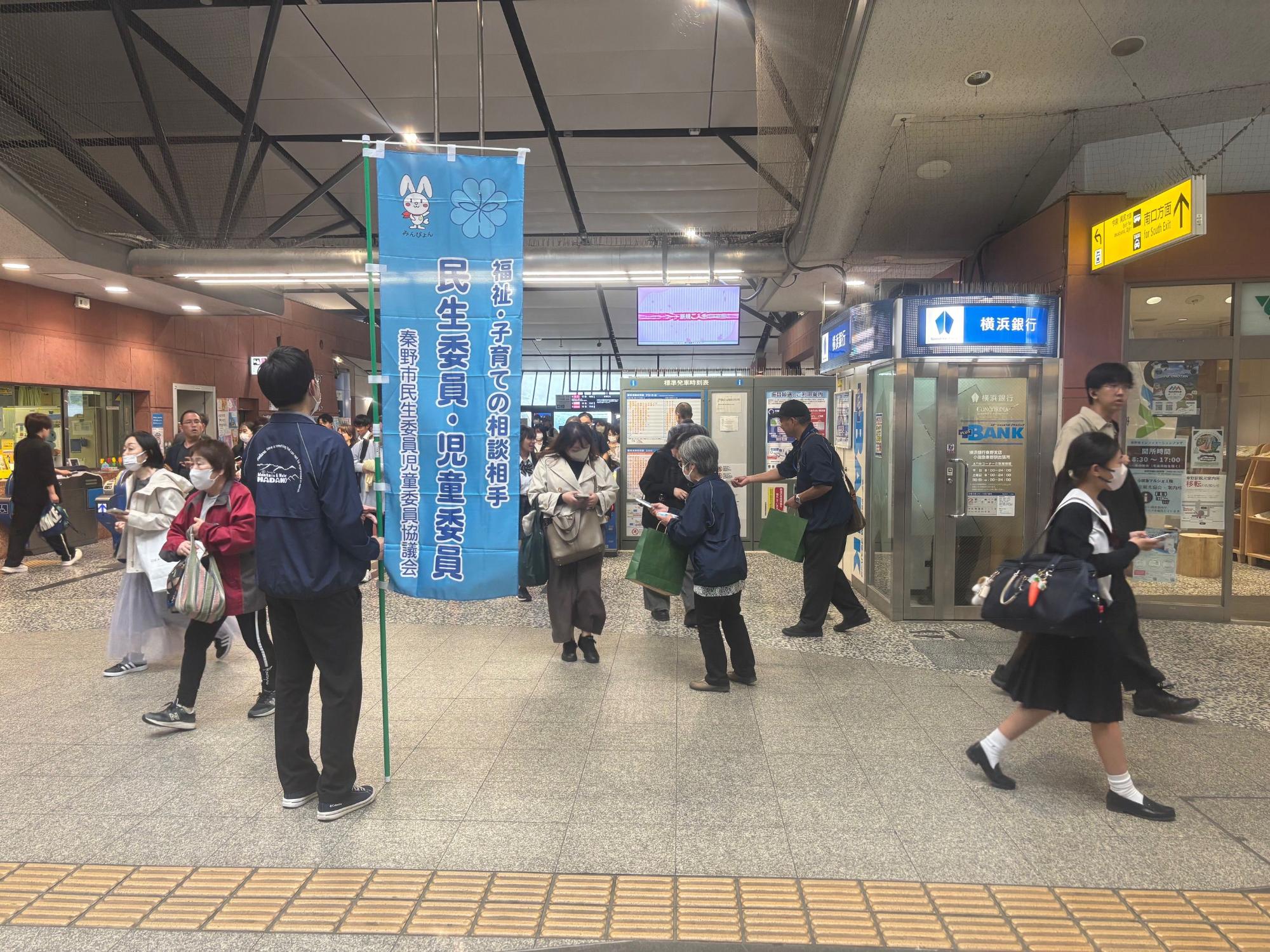 構内の通路で、「福祉・子育ての相談相手」と書かれたのぼり旗のそばに立つ若い男性が、行き交う人々にチラシを配っている風景を捉えた写真