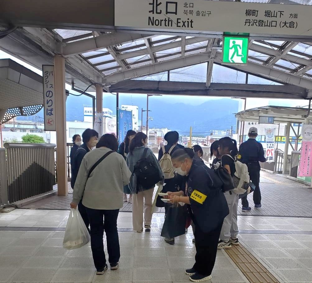 駅の改札前で、黄色い腕章をつけた複数の人々が、行き交う利用客にパンフレットやチラシを配っている写真