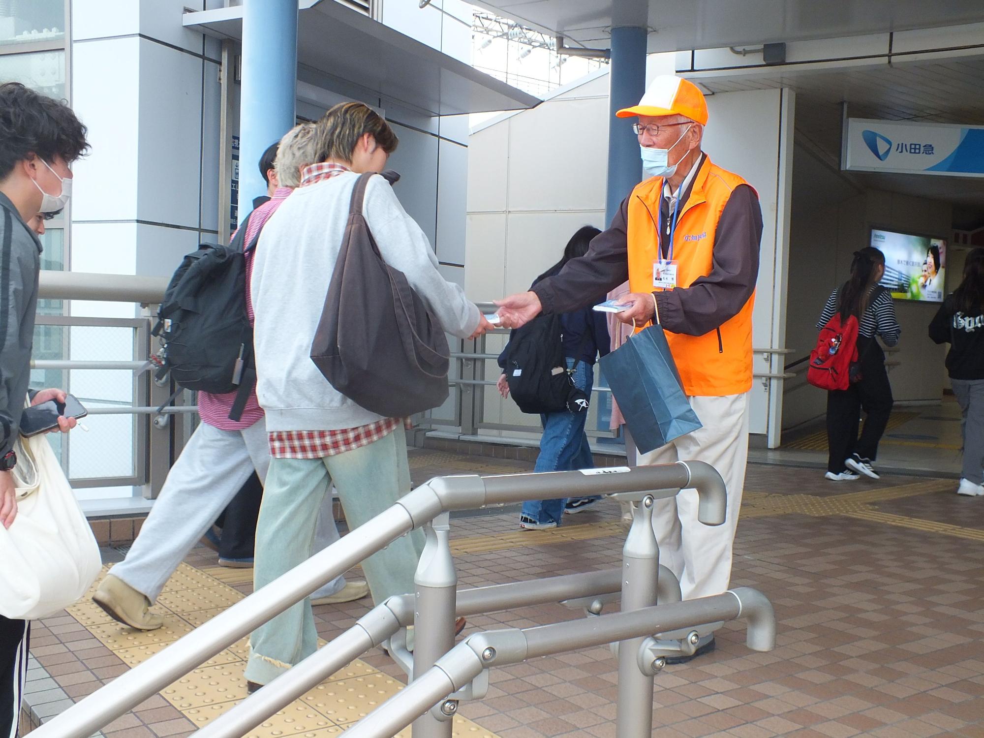 駅のロータリーでオレンジ色のベストと白い帽子を着用した男性が、通行人に青い袋を差し出して何かを配っている様子を捉えた写真