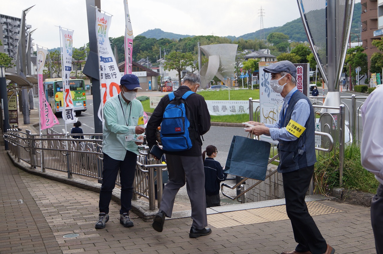 広報活動の一環として、駅前や道路の入り口でマスクを着用した2人の男性が、通行人に青い袋やチラシを手渡している写真