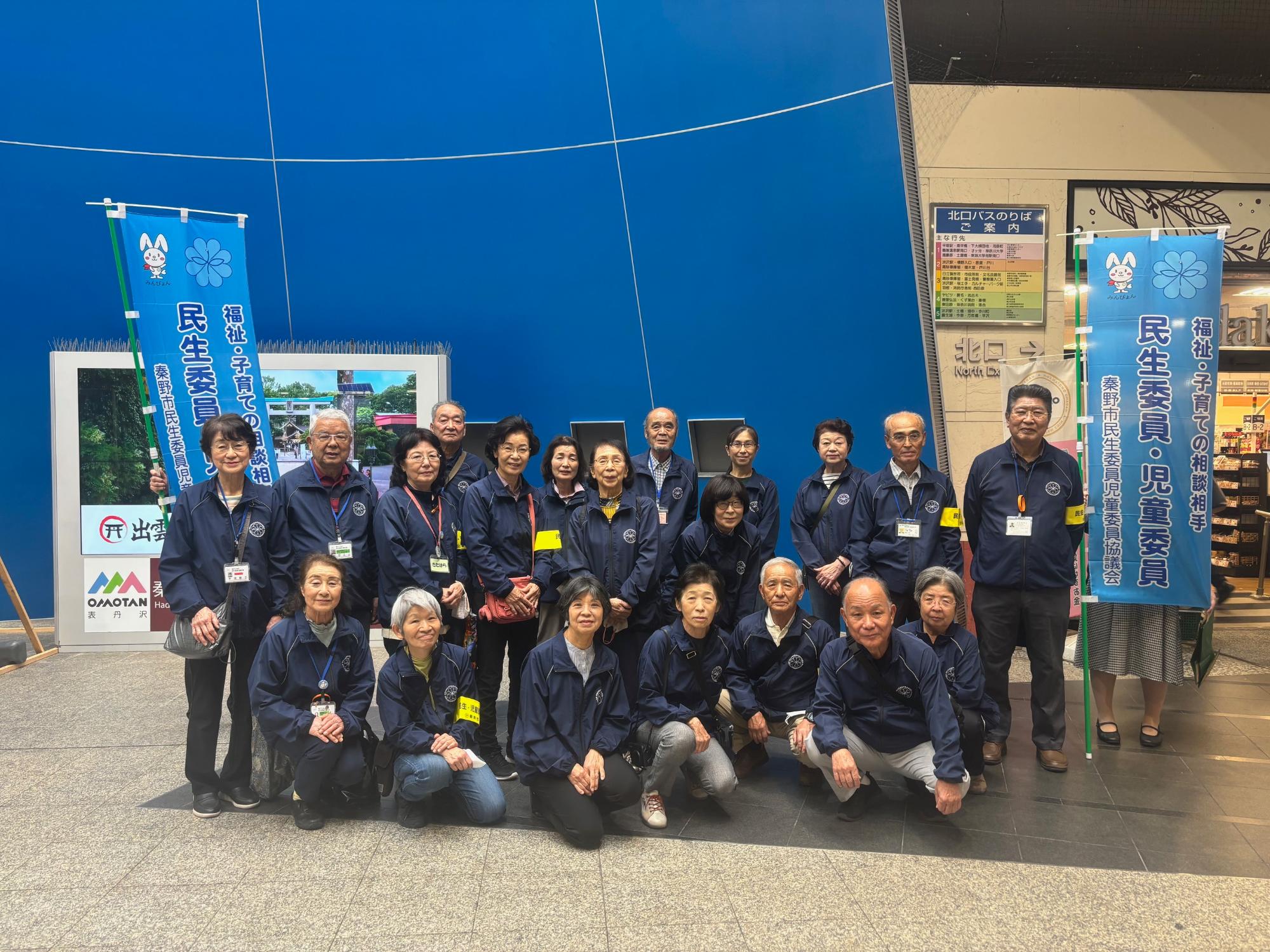 「民生委員・児童委員」と書かれたのぼり旗を持つ人を含め、青い上着を着用した男女が集合している写真