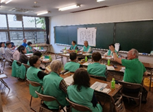 緑色のベストを着た大勢の人が、机に向かいながら話し合いを行っており、男性が手を挙げて発言している様子の写真