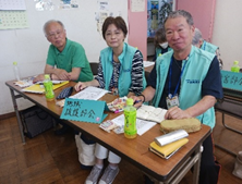 机に座った男女3人がカメラに向かって微笑みかけており、机の上には「地域援護部会」と書かれた札が置かれている写真