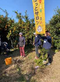 「ママとキッズのほっとサロン」と書かれた旗を持った男性が立つミカンの木が並ぶ果樹園で、子供を連れた親子が性と楽しそうにミカン狩りを楽しんでいる様子の写真