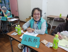 「広報部」と書かれた札が置かれた机に座る女性が、カメラに向かって満面の笑みを浮かべている写真