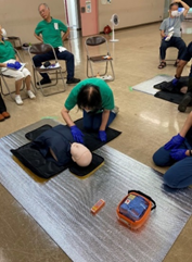 講習会場の床に横たわった心肺蘇生訓練用の人形に対し、緑色のベストを着用した女性が心臓マッサージの手順を実践しており、周囲の参加者たちがそれを見守る講習会の写真