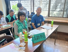 テーブルに置かれた「高齢部会」と書かれた札のそばで、高齢の男女2人がにこやかな表情で座っている写真