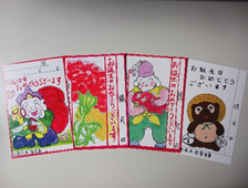 様々なデザインが描かれた4枚のメッセージカードが並んでおり、左から大黒様、赤い花、福の神、信楽焼のタヌキのイラストと「お誕生日おめでとうございます」などの文字が描かれている誕生日カードの写真