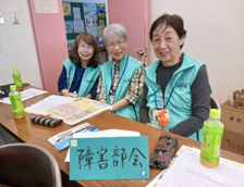 会議室の机に座った3人の女性がカメラに向かって微笑み、テーブルには「障害部会」と書かれた青い札が置かれている写真