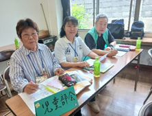 「児童部会」と書かれた札が置かれた机に座る3人の男女が、カメラに向かって優しく微笑みかけている写真