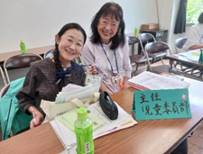 「主任 児童委員部」と書かれた札が置かれた机に座っている2人の女性が、カメラに向かって満面の笑みを浮かべている写真