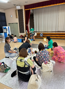 広い部屋の中で複数の母親たちが子供たちと一緒に床に座り、おもちゃや絵本を使って交流している様子を撮影した写真