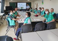 前方に設置されたスクリーンの傍に立つ男性と、長机を囲んで座っている緑色のベストを着た大勢の人々が、カメラを向いている研修の様子を捉えた写真