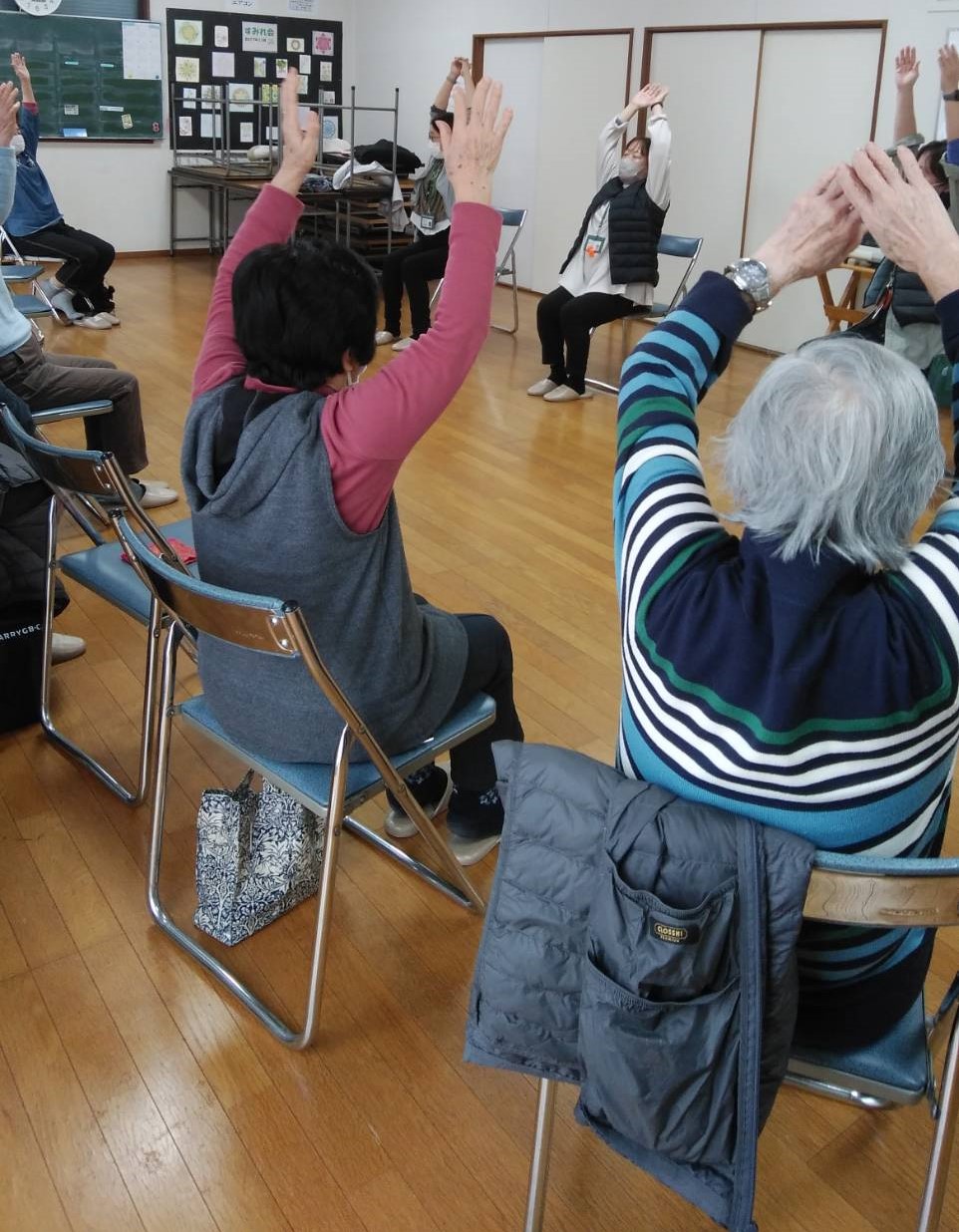 椅子に座った複数の高齢者が、両手を高く上げてさわやか体操をしている様子を後ろから撮影した、レクリエーション活動の写真