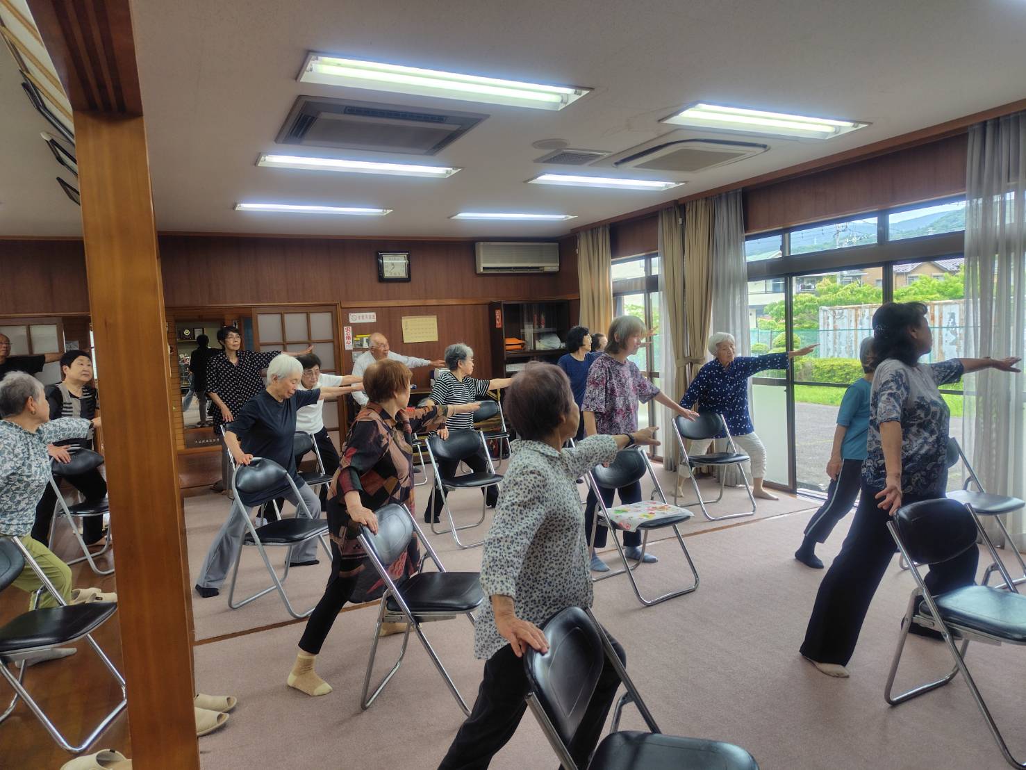 複数の高齢者が座っていた椅子の背を利用しながら、両足を前後に大きく広げて立ち、右手で椅子の背を持ち、左手を前に真っすぐ伸ばしてさわやか体操をしている様子を撮影した写真