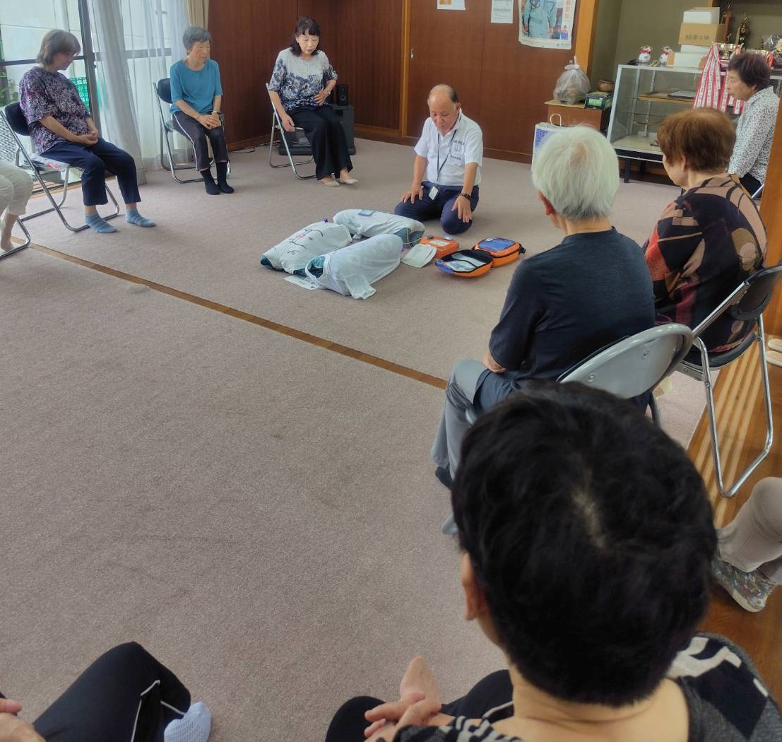 室内の端に広がって座っている数人の高齢者が座って見守る中、男性が床に置かれた人形を使って、AEDの使用方法について説明している様子を撮影した講習会の写真