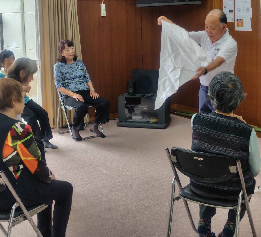 男性講師が広げた白い布を三角形に折って見せている様子を、周りに座っている複数の高齢者たちが真剣な表情で見ている、救命救急勉強会の様子の写真