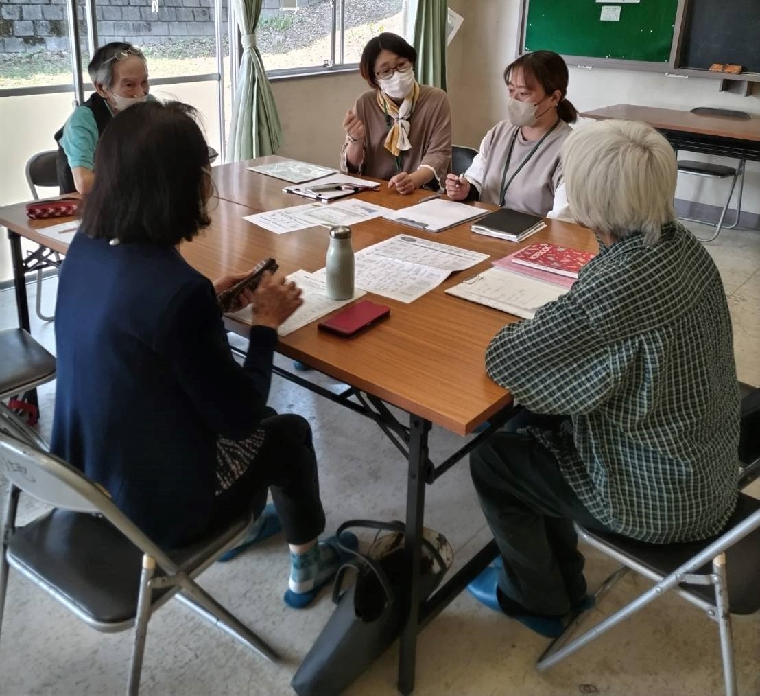 黒板が設置された室内で、2台の長方形のテーブルを囲んで座っているマスクを着用した5人の参加者が勉強会を行っている様子の写真