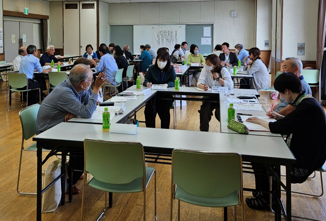 学校の教室や公民館のような明るい部屋で、多数の参加者がロの字型に並べられたテーブルを囲み、真剣に話し合ったり資料にメモを取ったりしている意見交換の写真