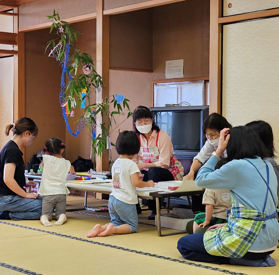 畳の部屋に七夕の飾りがついた笹があり、大人と子どもたちが低い机を囲み、絵を描いたり工作をしたりしている様子の写真