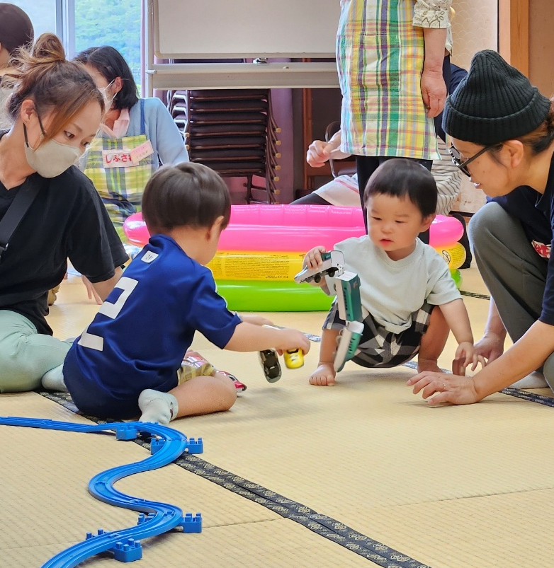 2人の子どもが玩具の車で遊んでおり、近くで大人が見守っている様子の写真