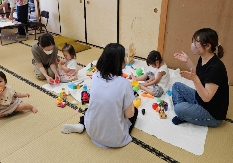 畳の部屋で、複数の子どもが大人と一緒におもちゃで遊びながら交流している様子を写した、地域の子育て支援活動の写真