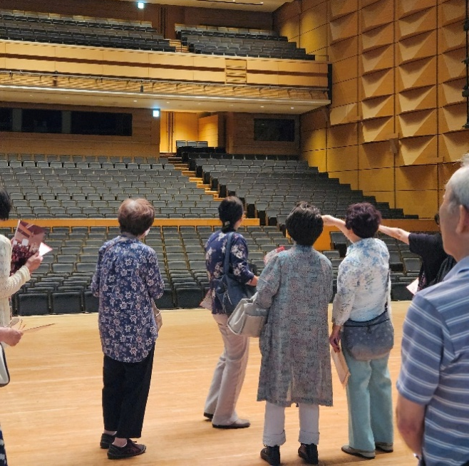 舞台上で客席側を向き、背の高い木目調の壁と黒い座席が並ぶ階段状の大ホールを見学する数人の参加者の写真