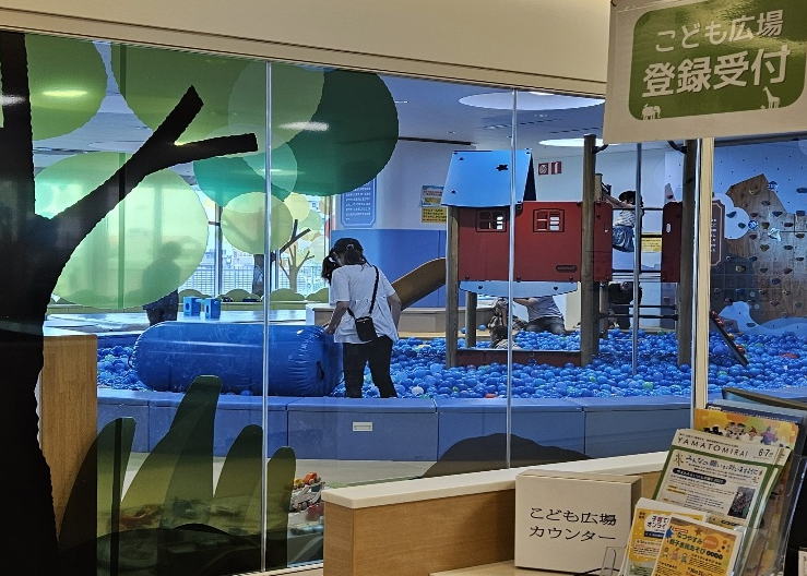 透明なガラス窓越しに、青いボールプールの中にいる子どもたちが、赤い遊具や大きな青いエア遊具で遊んでいる屋内施設と受付カウンターの景色の写真