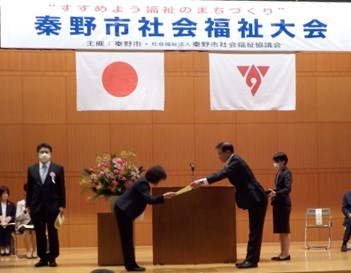 「秦野市社会福祉大会」と書かれた横断幕が掲げられたステージ上で、檀上前でスーツ姿の男性と女性が向かい合い賞状を手渡している様子の写真