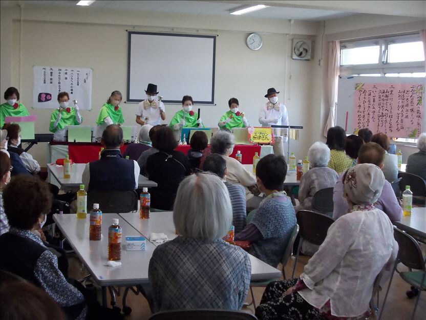地域の公民館のような会場で、緑と白の衣装を着た7人の奏者がハンドベルを演奏し、テーブルに着席した大勢の人達が演奏を楽しんでいる様子を捉えた写真