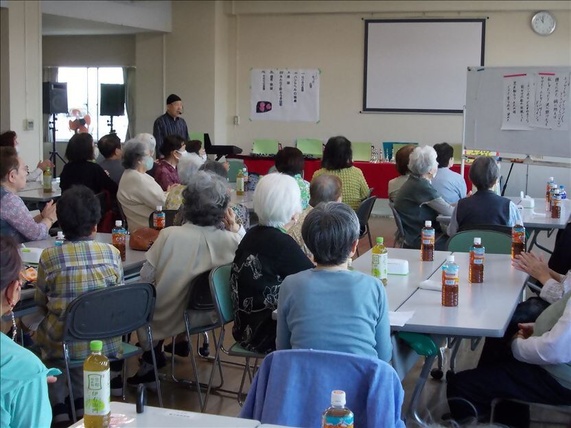 広い室内で、ペットボトルのお茶を前に置いたテーブルと椅子に着席した大勢の高齢者が、黒い帽子と縞模様のシャツを着た男性が歌っている内容に耳を傾けている様子の写真