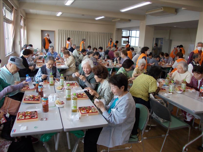 地域コミュニティセンターのような広い室内で、大勢の高齢者がテーブルを囲んで座り、お茶のペットボトルと仕出し弁当のような食事を楽しみながら交流している昼食会の風景写真