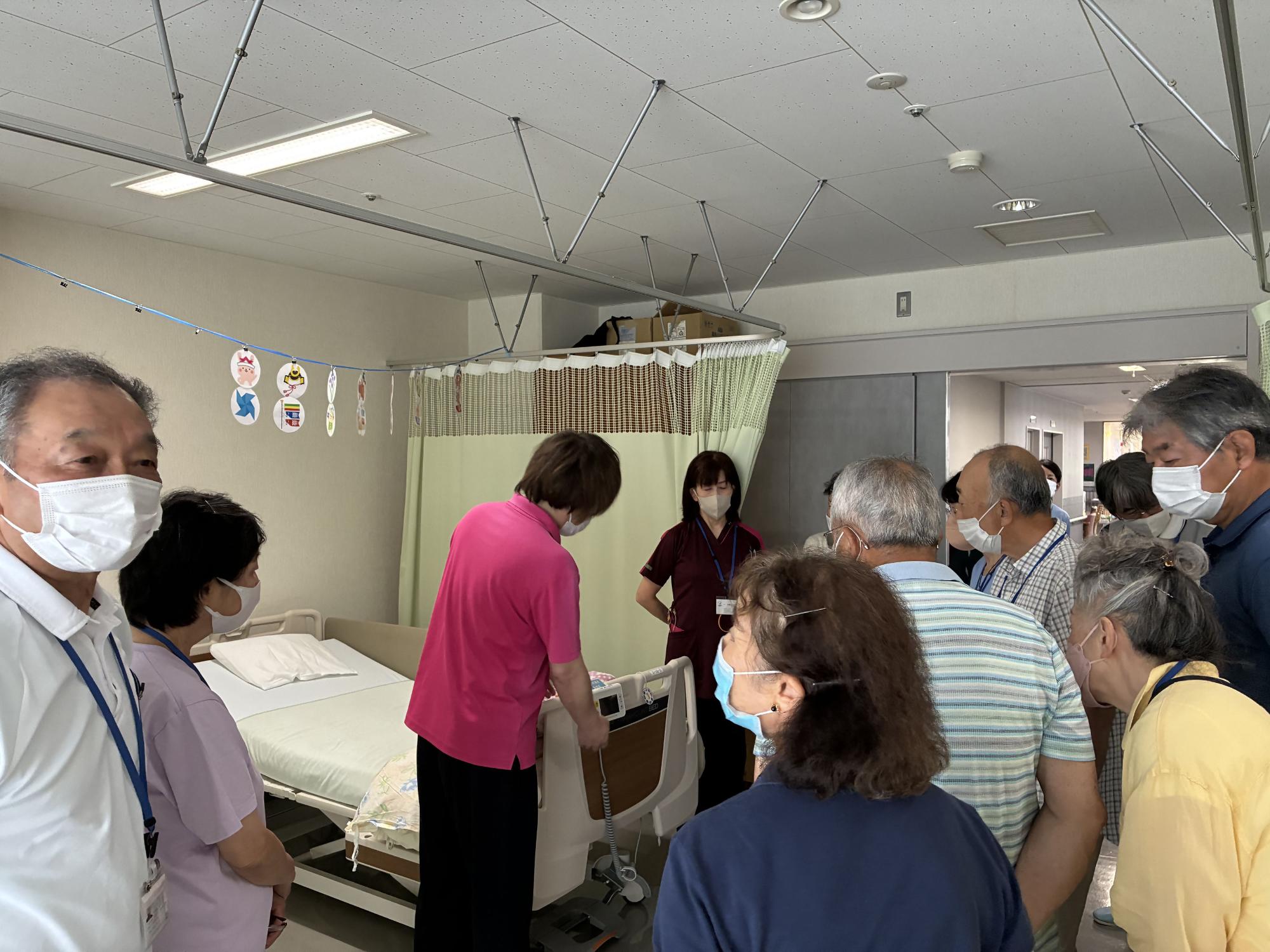 病院のベッドがある室内で、数人の高齢者が介護ベッドの操作方法について、介護士や看護師から説明を受けている写真