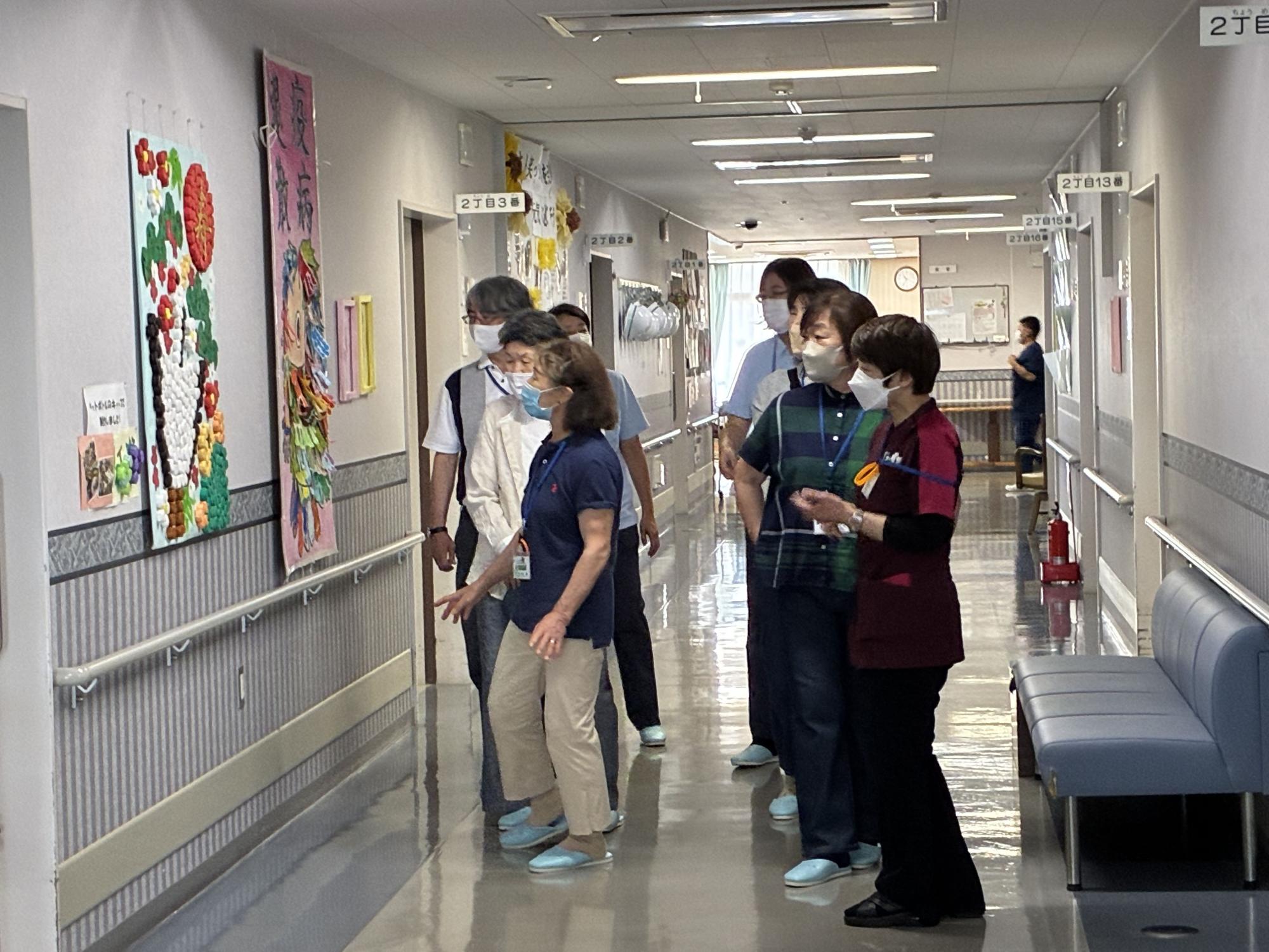 病院の長い廊下で、数人の高齢者が介護士や看護師と一緒に、壁に飾られている手作りの色鮮やかな壁飾りの作品を鑑賞している写真