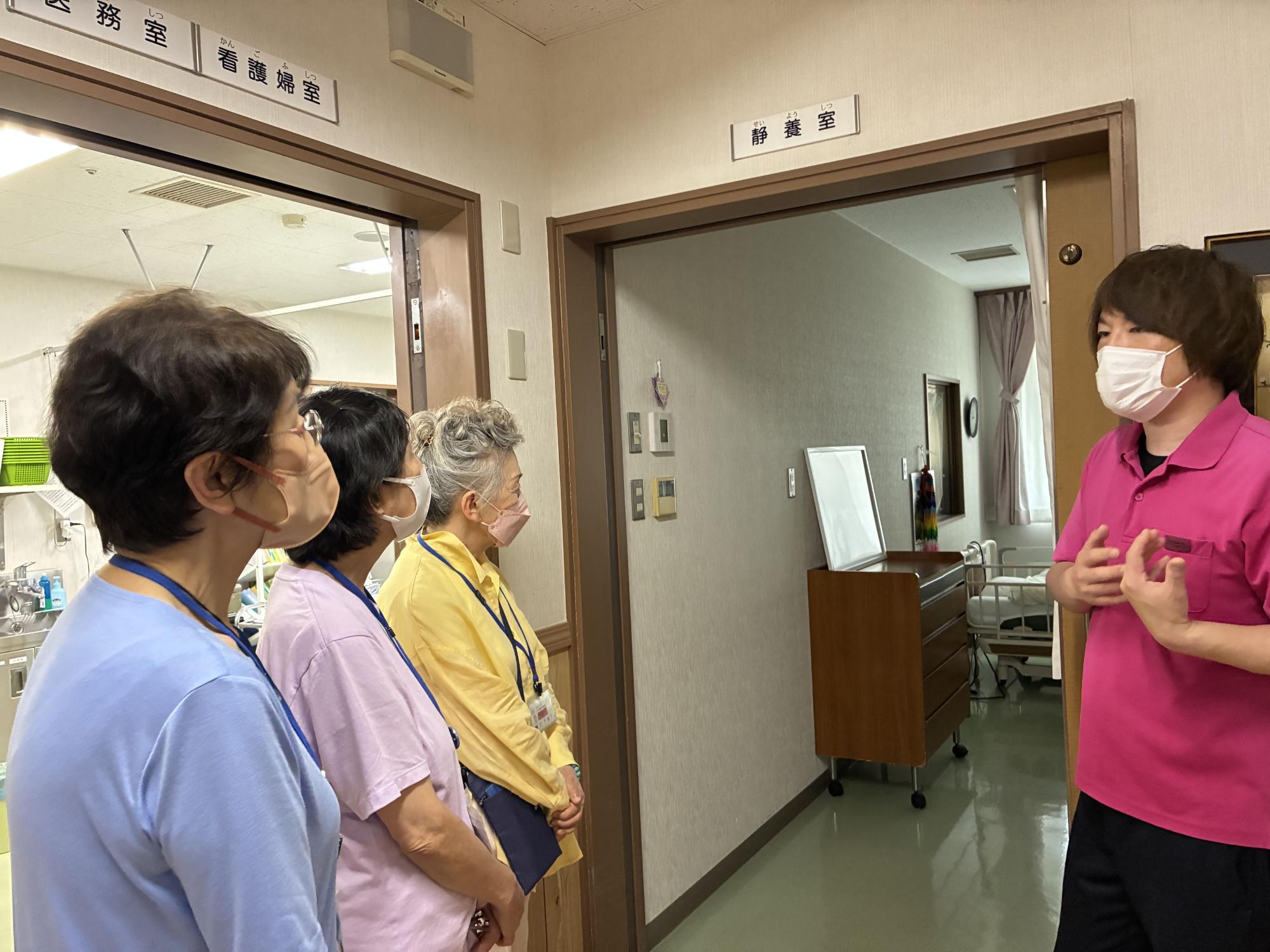 病室の前で、数人の高齢者が介護士や看護師から、病室内の設備や概要について説明を受けている様子の写真