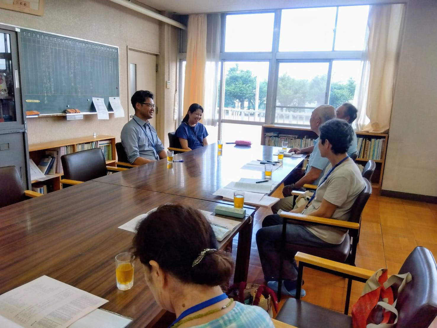 複数の大人が長いテーブルを囲んで座っており、窓から明るい光が差し込む部屋で、飲み物を飲みながら談笑している懇談会の様子の写真