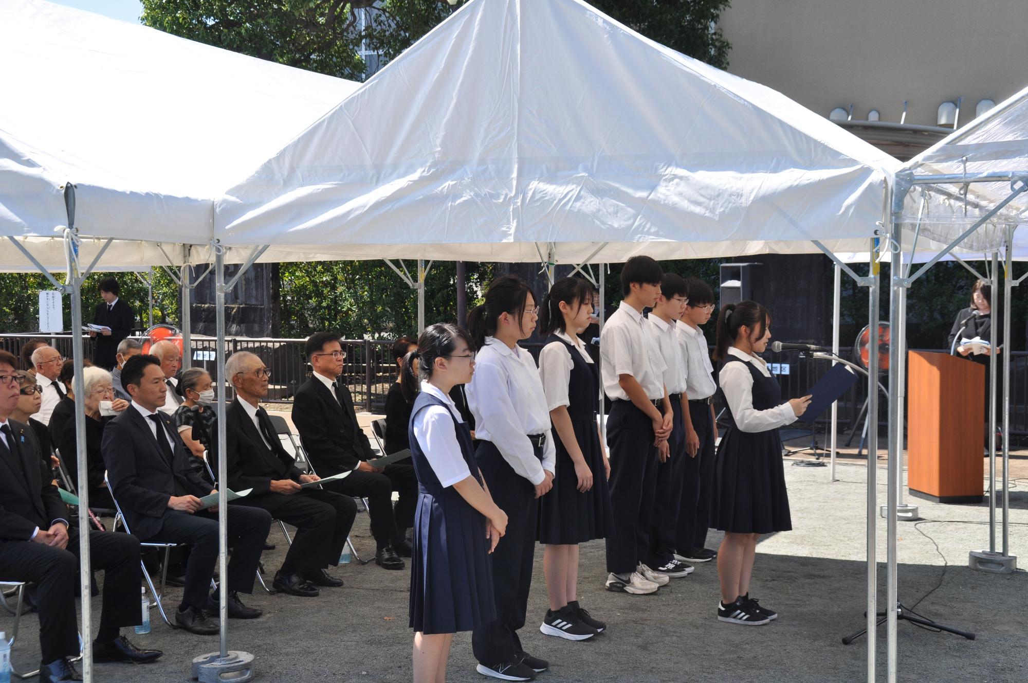 白いテントの下で制服を着た複数の学生たちが横一列に並び、代表の女子生徒がマイクに向かって立っており、黒いスーツ姿の多くの人々が静かに座って聞いている、式典の様子を写した写真