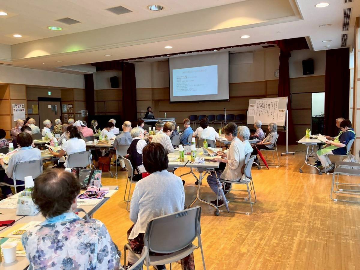 会場前方に資料が映し出されたスクリーンが設置され、多くの高齢者がテーブルと椅子が並んだ部屋で、檀上にいる講師の話を聞いている様子を撮影した写真