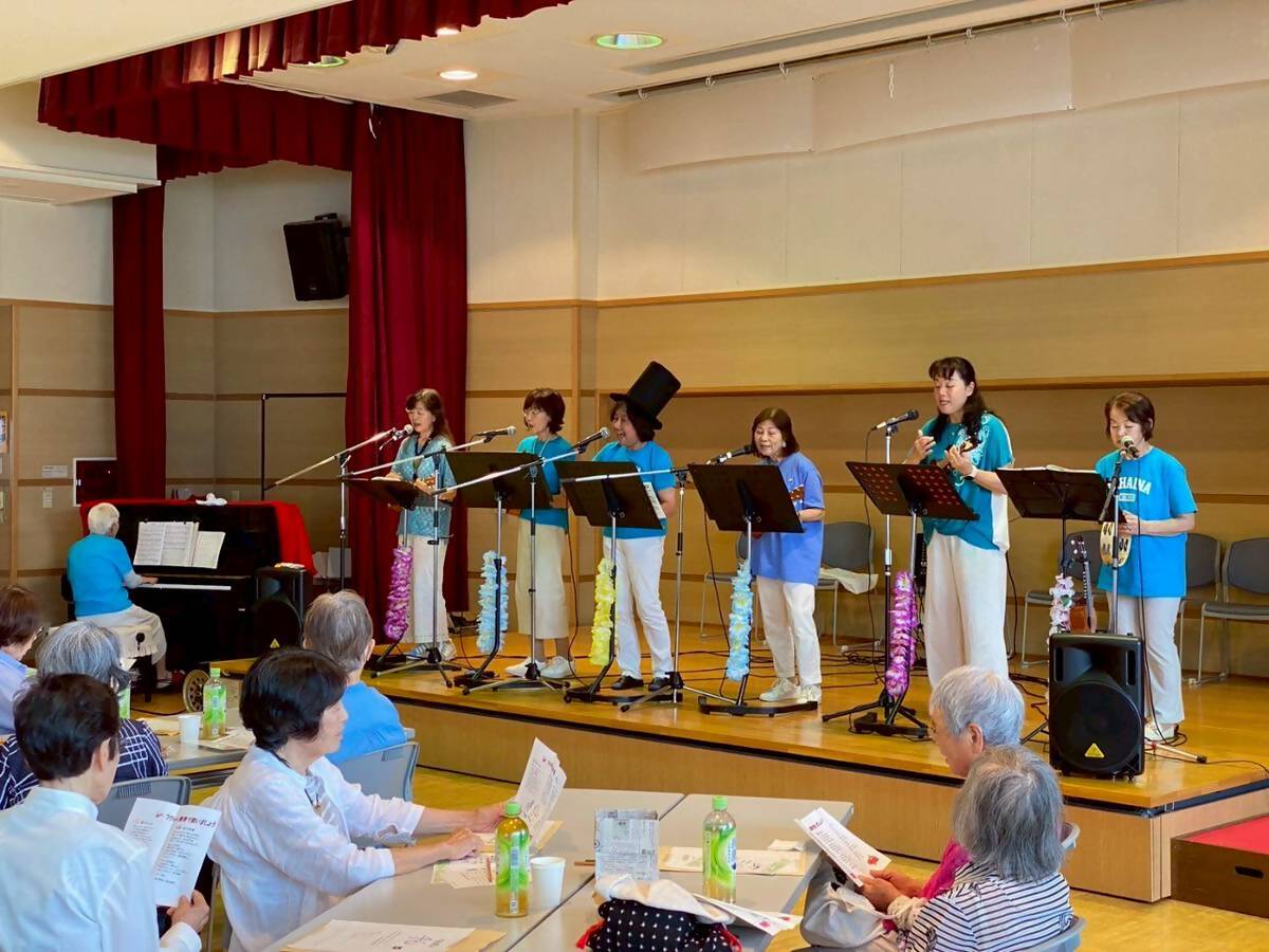 ピアノの演奏にあわせて水色のシャツを着たステージ上の6人の女性が、ウクレレを弾きながら歌を披露し、テーブルの席に座った参加者が熱心に聞いている様子を撮影した写真