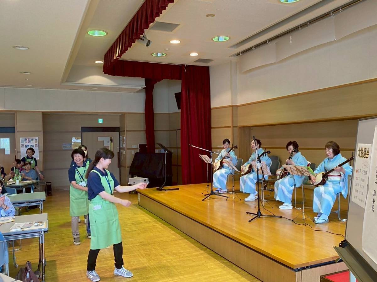 ステージ上の椅子に座った水色と白のラインが入った着物を着た4人の女性が演奏する三味線の音色に合わせて、お揃いの薄緑色のエプロンを着用した3人の女性スタッフがステージ下でパフォーマンスをしており、多くの観客が座って手拍子をしながら見ている写真