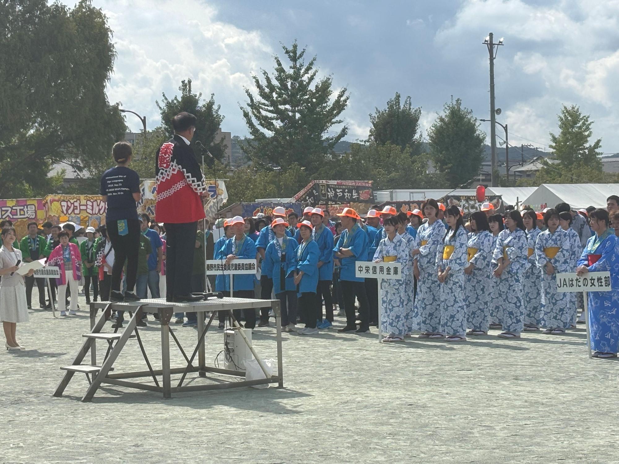 屋外イベント会場で青や緑の法被や浴衣を着た複数の地域団体が団体名プラカードを掲げて整列し、檀上の法被を着た男性がマイクで開会挨拶を行う地域祭りの写真