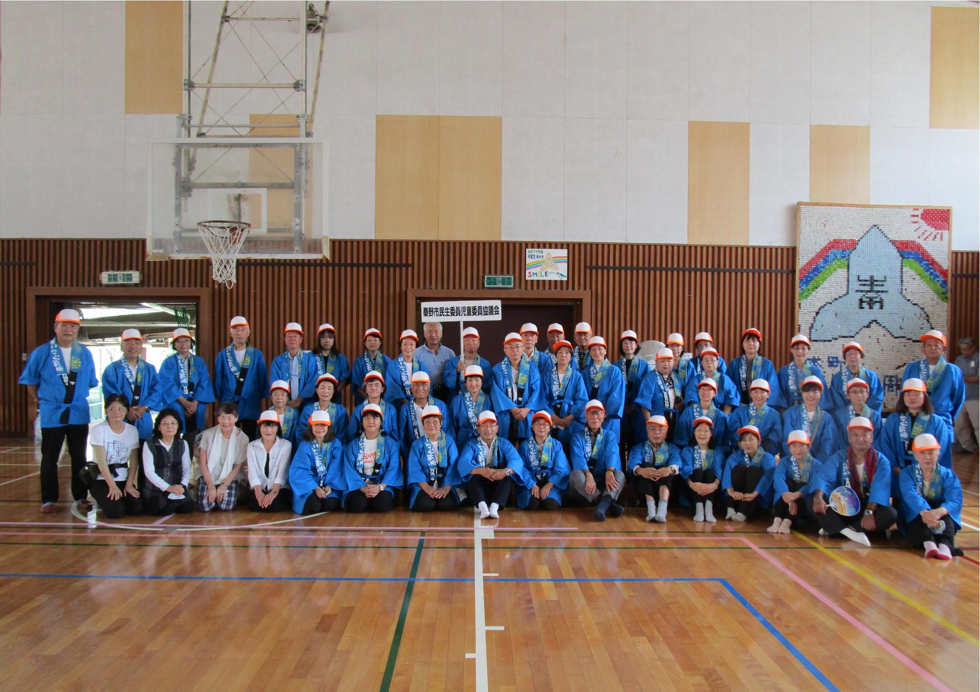 体育館のバスケットコートで、青い法被とオレンジの帽子を着用した数十名のグループが、前列は床に座り、後列は立ち上がって、和やかな雰囲気で記念撮影に応じる集合写真